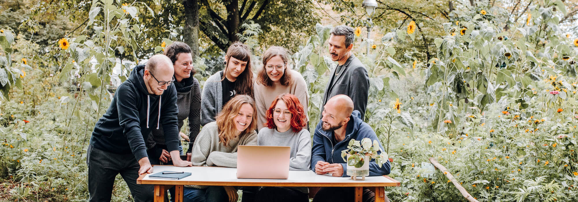 Ein Teambild eurer Lieblings-NGO-Agentur. Acht Personen sitzen und stehen mitten im Grünen um einen Schreibtisch mit einem laptop und einer netten Glückspflanze darauf. Alle schauen in den Laptop und lächeln.