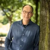 Mittelalter Mann mit Brille, kurzen Haaren und einem dunkelgrauen Hemd lehnt lässig an einem Baum.