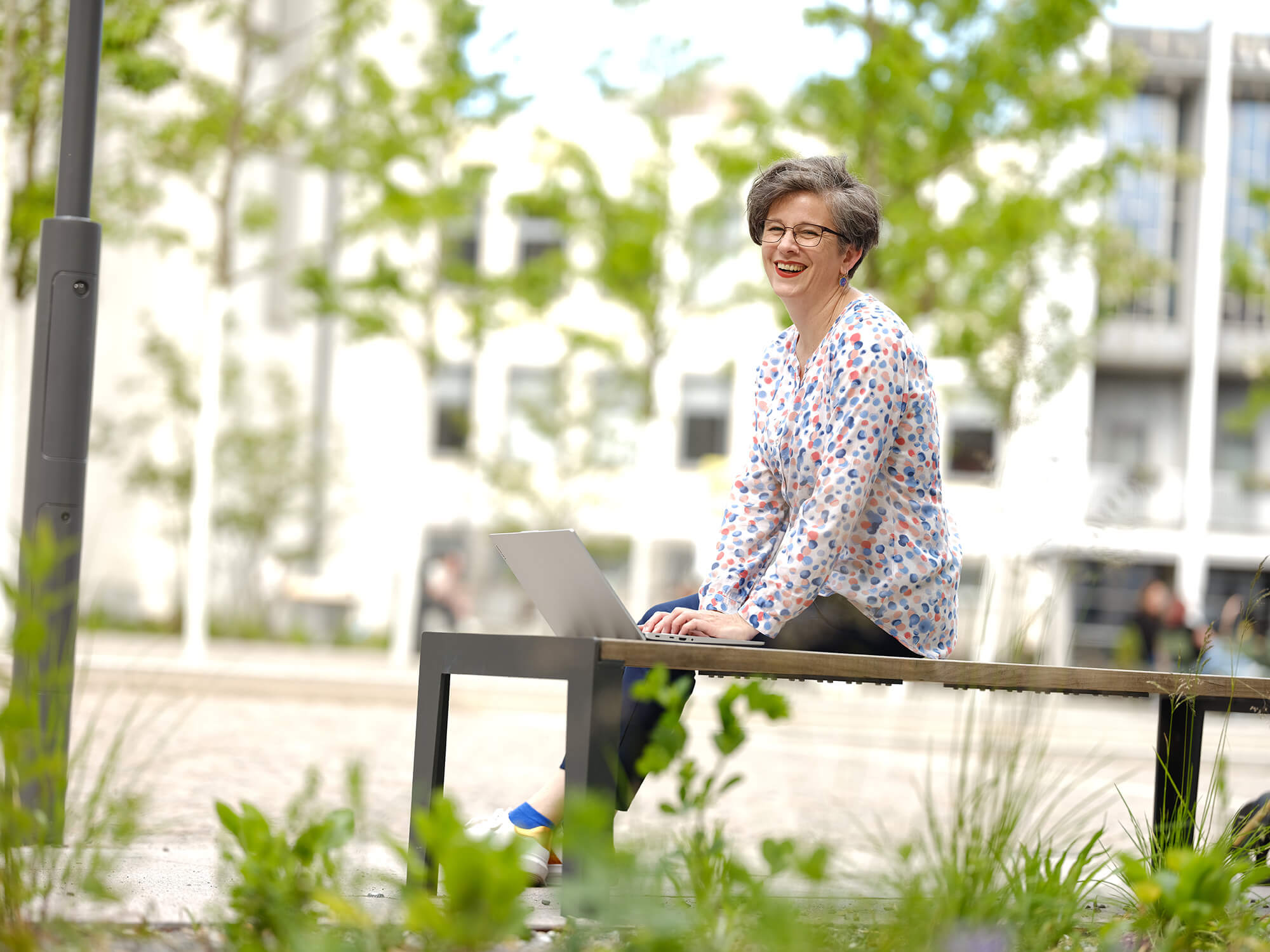 Eine Frau mit kurzen Haaren sitz auf einer Bank mit aufgeklapptem Laptop vor ihr. Sie lächelt und schaut in die Ferne. 