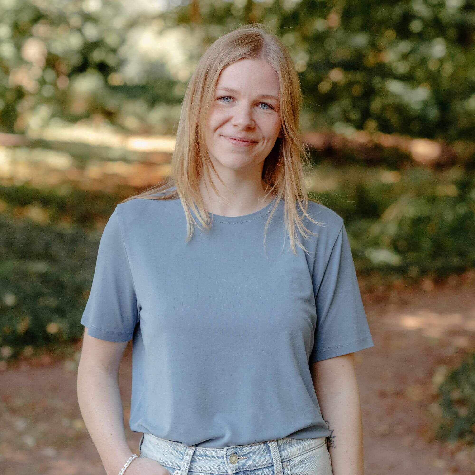 Frau in hellblauem T-Shirt steht draußen im Grünen. Sie hat mittellange, blonde Haare und lächelt.
