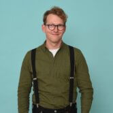 Mann mit Brille, orangen Haaren, dunkelgrünem Hemd und schwarzen Hosenträgern.