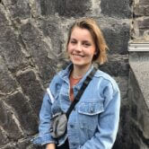 Frau mit kurzen Haaren und Jeansjacke. Lächelt und steht vor einer Steinmauer.