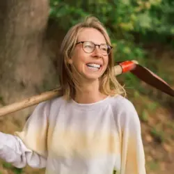 Frau mit Brille lächelt und hat einen Spaten geschultert.