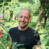 Mann mit sehr kurzen Haaren, lächelt und steht in einem dicht bewachsenen Garten.