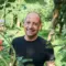 Mann mit sehr kurzen Haaren, lächelt und steht in einem dicht bewachsenen Garten.
