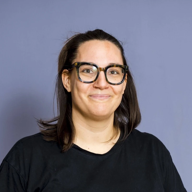 Person mit schulterlangen, dunkelbraunen Haaren, großer Brille, im schwarzen Pullover. Lächelt.